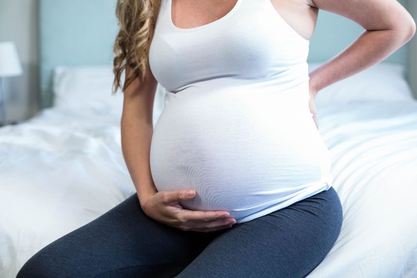 A pregnant woman sitting on a bed, holding her abdomen with one hand and supporting her back with the other, depicting the physical strain that can lead to Diastasis Rectus Abdominis during or after pregnancy.